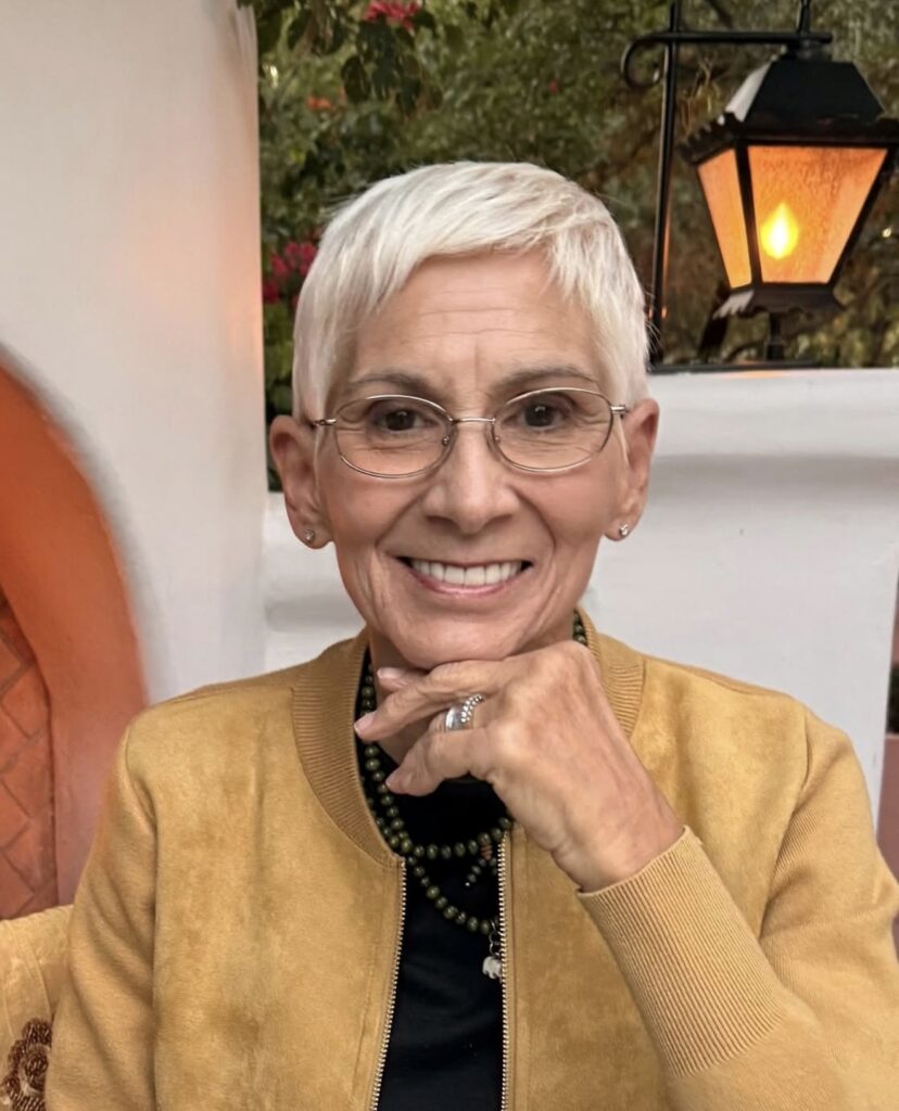 Smiling older woman with short gray hair and glasses rests her chin on her hand, wearing a yellow jacket and layered necklaces. A lantern and greenery in the background add warmth.