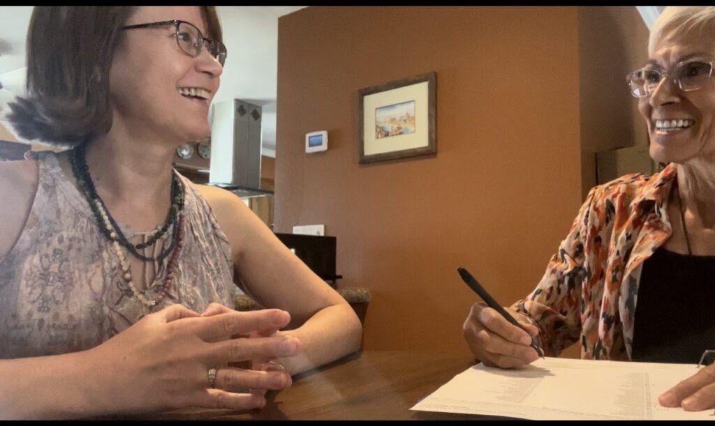 Two women engage in a lively conversation at a table, with one writing on notepad. The setting is warmly decorated, suggesting a casual meeting.
