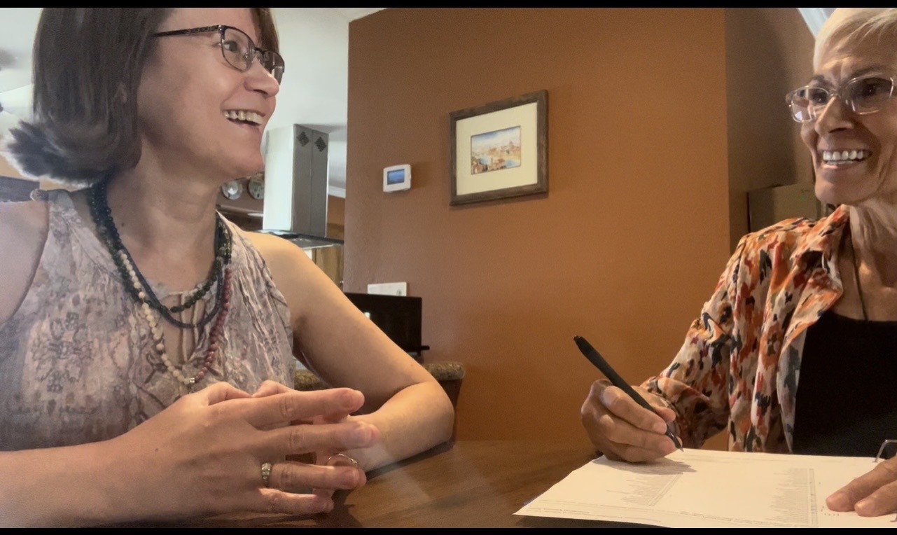 Two women engage in a lively conversation at a table, with one writing on notepad. The setting is warmly decorated, suggesting a casual meeting.